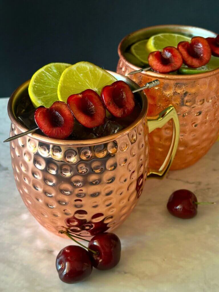 Two copper mugs full of tart cherry mule and garnished with limes and pitted cherries
