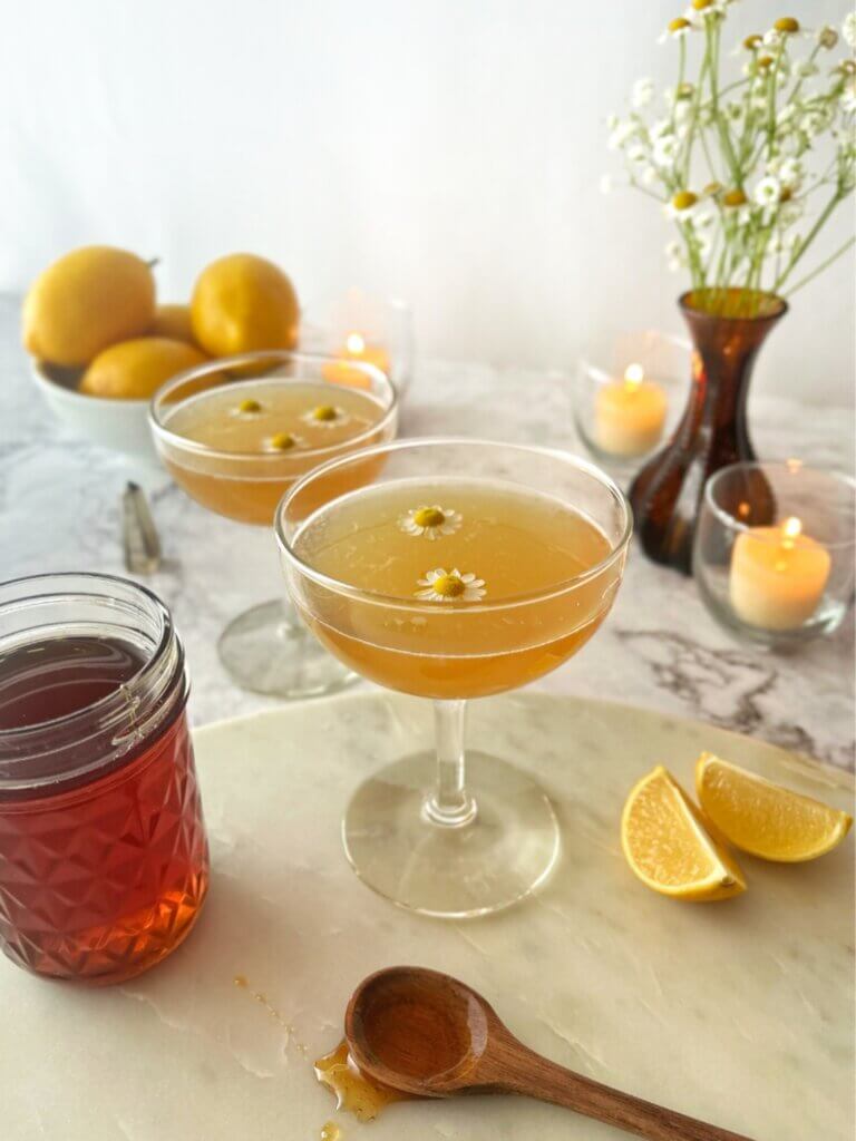 A bowl of lemons, a mason jar of honey and a vase of chamomile hints at the chamomile drink recipes served in two delicate coup glasses on a marble countertop