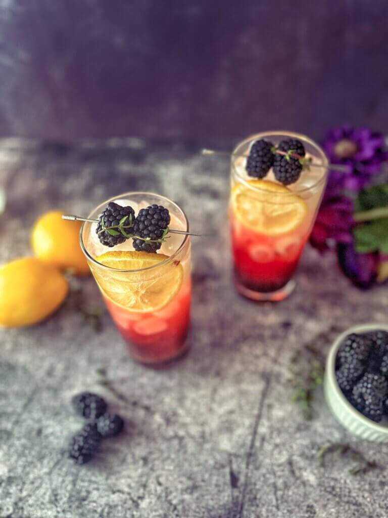 Two blackberry-thyme tom collins mocktails topped with a skewer of blackberries wrapped with a sprig of thyme.