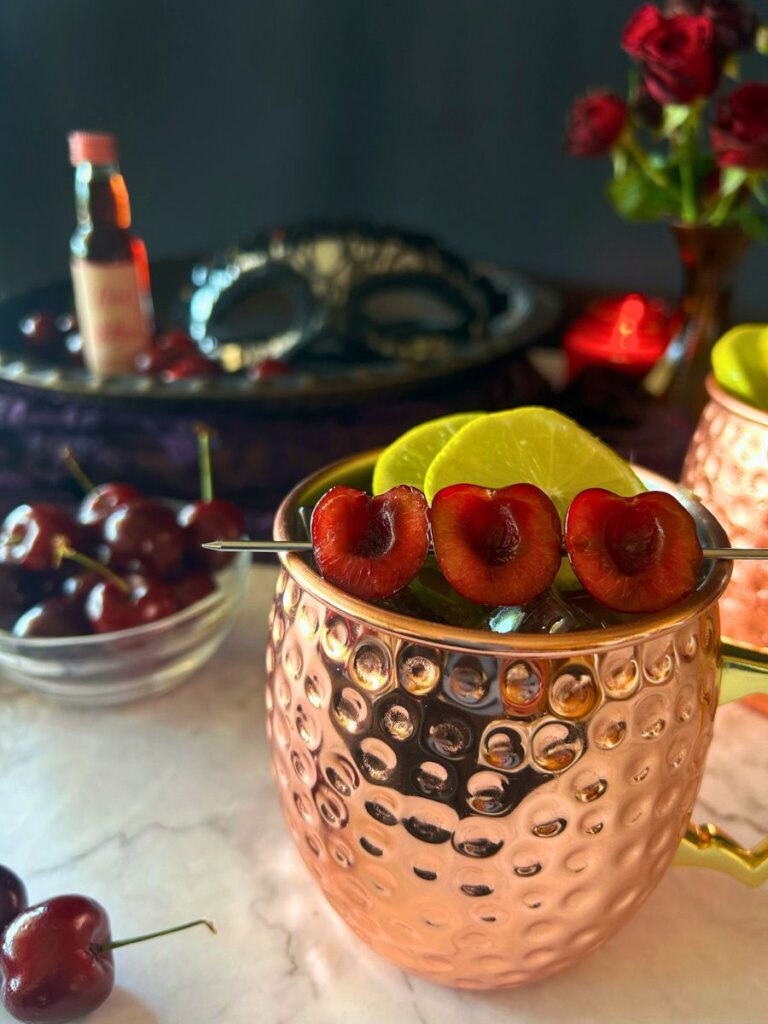 Tart cherry mules surrounded by love bitter, a bowl of cherries, a red candle and a vase of tea roses