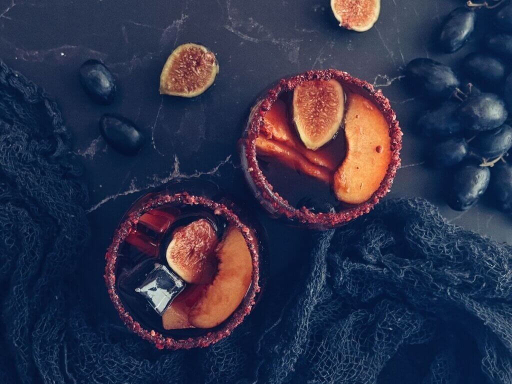 Two glasses of Halloween sangria surrounded by assorted fruit garnishes like black figs and grapes