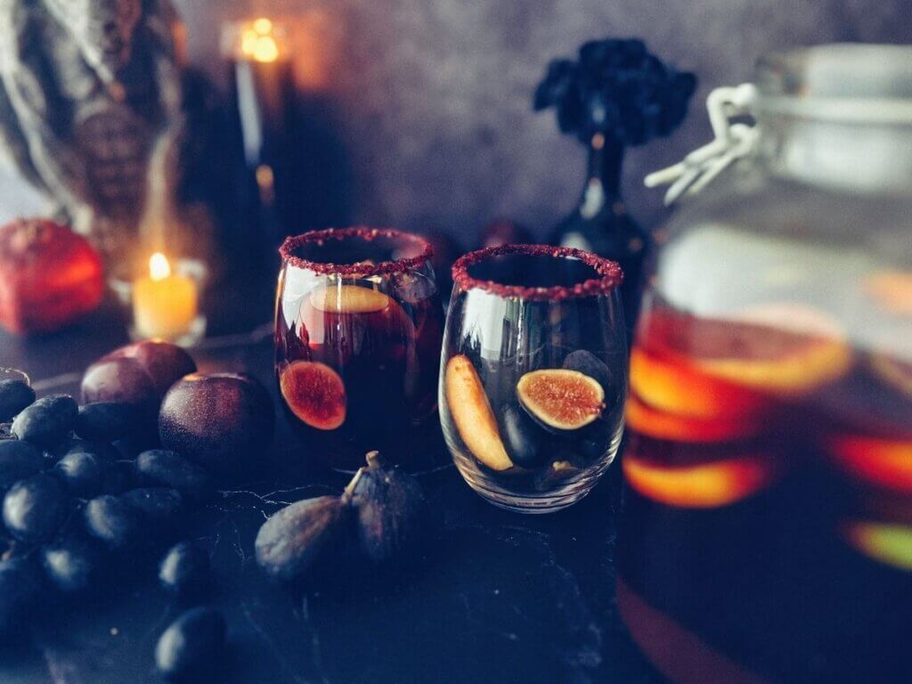 Two glasses of black sangria surrounded by assorted fruit garnishes like black figs, grapes, plums and pomegranate