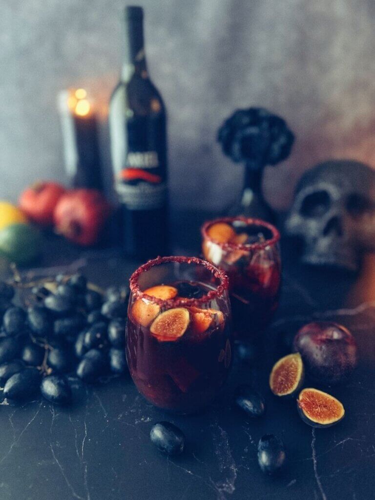 Two glasses of fall sangria mocktail surrounded by assorted fruit garnishes and spooky decor like a vase with black flowers and a black skull